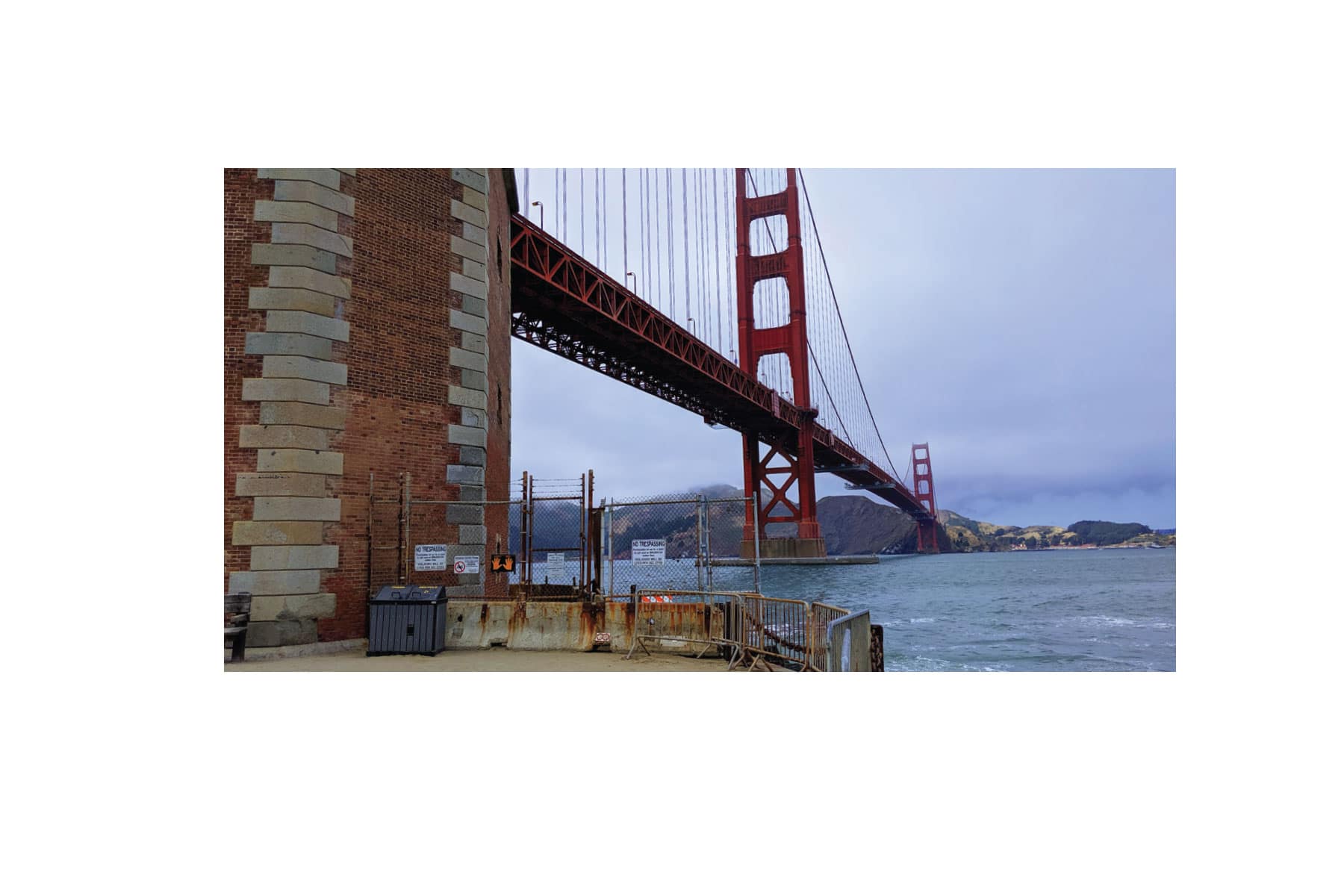 An image of the Golden Gate Bridge from the pier below. A fence with warning signs prevents approach.